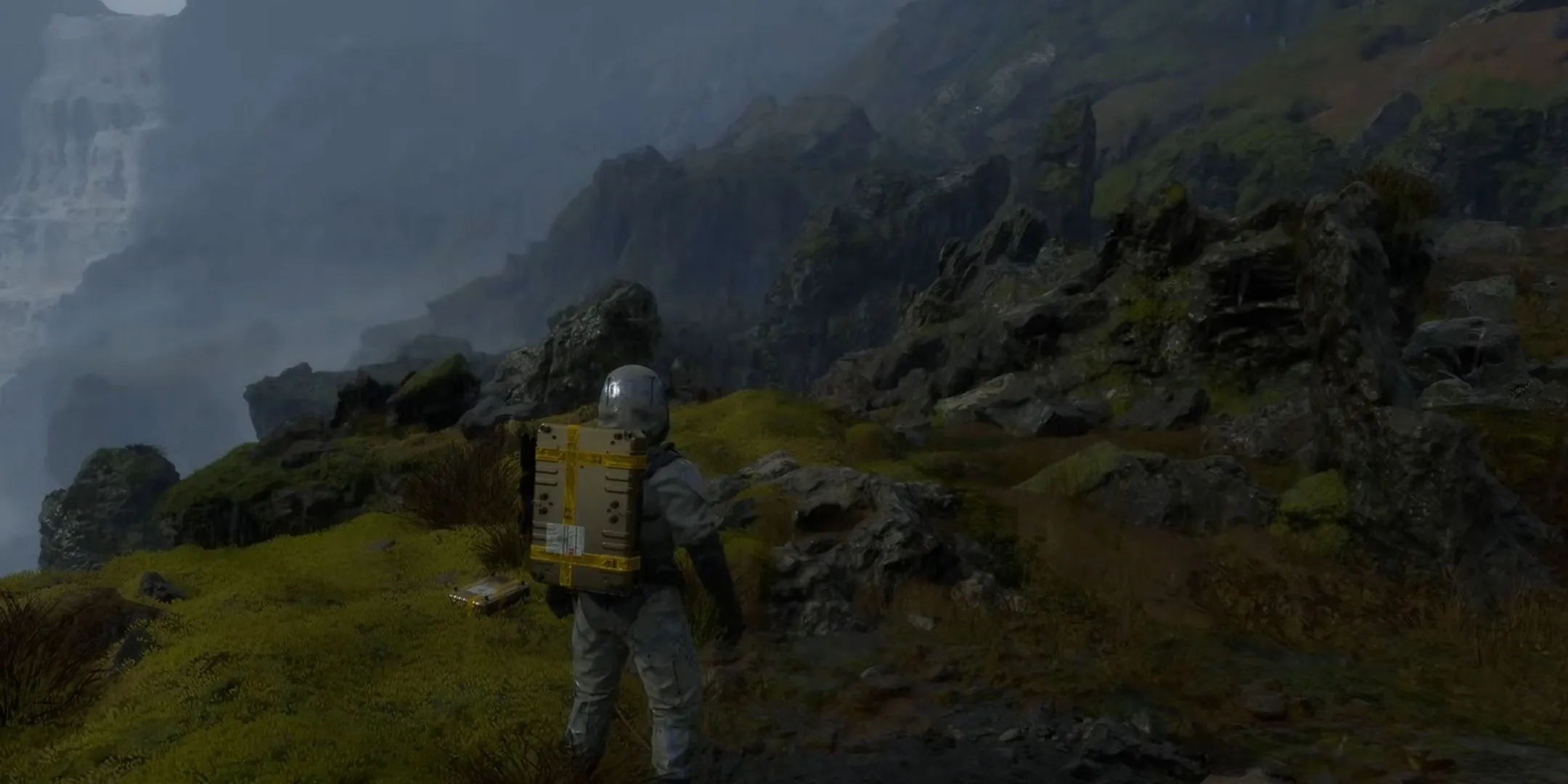 Sam Porter Bridges đang băng qua địa hình hiểm trở trong Death Stranding với các kiện hàng lớn sau lưng.